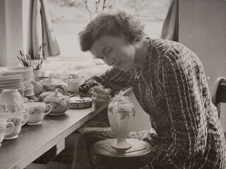 Black and white photograph of woman sat at a bench hand painting ceramics